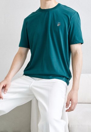 Teal cotton T-shirt with short sleeves, round neck, and a small multi-colored embroidered logo on the chest. Worn with white pants.