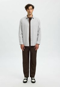 Gestreepte button-up shirt in lichtgrijs en wit, gelaagd over een donkerbruine t-shirt, gecombineerd met rechte bruine broeken en zwarte schoenen met witte accenten.