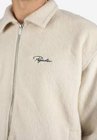 Cream-colored fleece jacket with a soft texture, zip closure, and a collar. Features black "Pegador" embroidery on the chest.