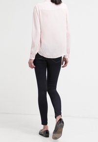 Blouse rose pâle à manches longues avec col, associée à un jean skinny foncé et des ballerines noires. La blouse présente une texture lisse et douce.