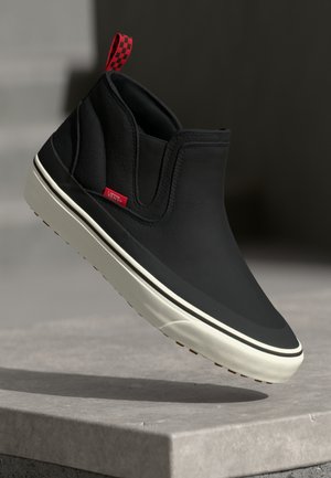 Black slip-on shoe with a textured finish, white sole, red checkerboard pull tab, and a small red logo on the side.