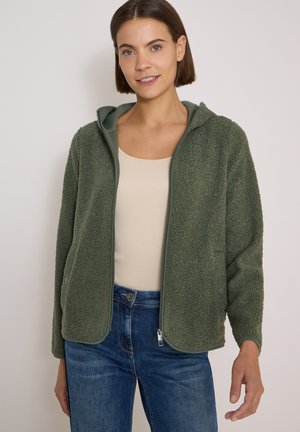 Kvinde iført en grøn fleecejakke med lynlås over en beige top og blå jeans, stående mod en ensfarvet lys baggrund.