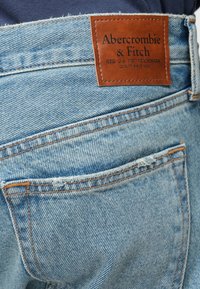 Svetlo modré džínsy s opotrebovanou textúrou. Obsahujú hnedý kožený štítok na páse s embosovaným nápisom "Abercrombie & Fitch".