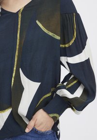 Blusa blu navy con pattern astratti bianchi e dorati, caratterizzata da maniche lunghe e voluminose e scollatura a V. Tessuto liscio e leggero.