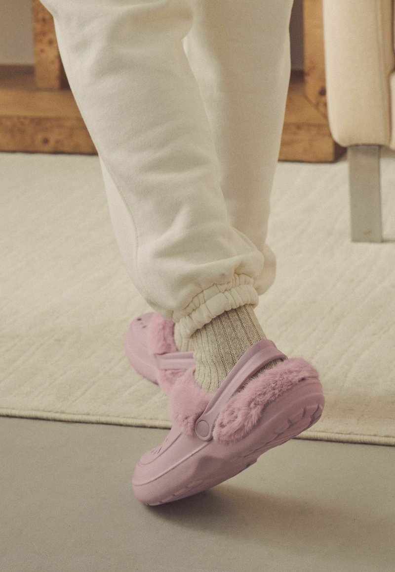Roze klompen met een fluffy rand, voorzien van een zachte textuur en een gevormd ontwerp. Draagt een lichtgrijs gebreid paar sokken en ontspannen witte broek.