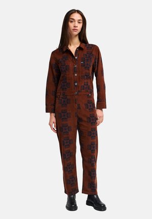 Jeune femme debout dans une combinaison marron à boutons avec des motifs géométriques foncés et des bottines noires, sur un fond clair uni.