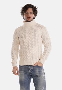 Maglione a collo alto lavorato a trecce di colore crema, con una consistenza spessa, polsini e orlo a coste, abbinato a jeans blu strappati.