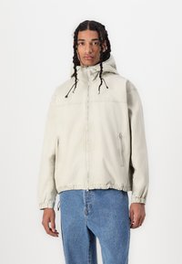 Light beige zip-up jacket with a hood, elastic cuffs and hem, featuring a front zipper pocket and minimal branding on the chest.