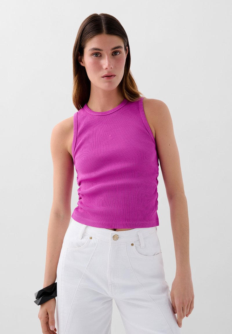 Top smanicato rosa a costine, aderente, con scollatura rotonda e arricciature laterali, abbinato a jeans bianchi a vita alta. Accessorio per capelli neri visibile.
