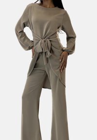 Completo beige in due pezzi composto da top a maniche lunghe con dettaglio nodo frontale e pantaloni a gamba larga, realizzato in tessuto liscio e leggero.