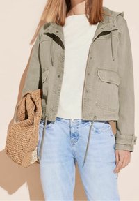Femme portant une veste vert clair sur un haut blanc et un jean bleu clair, tenant un grand sac en toile beige tissé à ses côtés.
