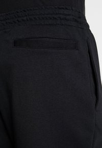 Mustat collegehousut, joissa on joustava vyötärönauha ja takatasku. Kangas on sileä ja hieman teksturoitu, suunniteltu mukavuuteen ja käytännöllisyyteen.