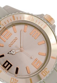 Montre ronde grise avec un boîtier en métal rose doré, marqueurs d'heure contrastants et un guichet de date à 6 heures. Bracelet en silicone lisse.