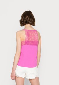 Top fucsia senza maniche con schiena in pizzo e texture liscia; indossato con pantaloni corti bianchi sfrangiati. Il top ha una forma aderente e una scollatura a barca.