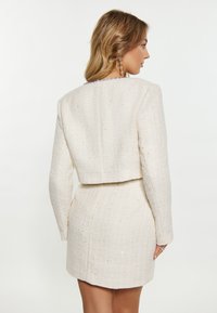 Rochie albă din tweed cu o jachetă scurtă, având mâneci lungi, material texturat și accente subtile strălucitoare pe suprafață.