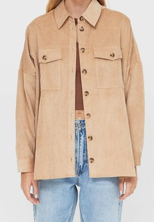 Beige corduroy blouse met knopen en borstzakken, open gedragen over een bruine cropped top, gecombineerd met lichtblauwe, hoog getailleerde jeans.