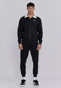 Veste de survêtement noire zippée avec un col et des rayures latérales de couleur crème, associée à un bas de jogging noir assorti. Détails du logo sur la veste.
