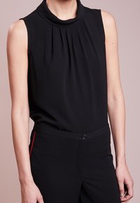 Svart ärmlös topp med hög krage, med veck av tyg vid bröstet. I kombination med svarta byxor, som har en röd sidolinje.