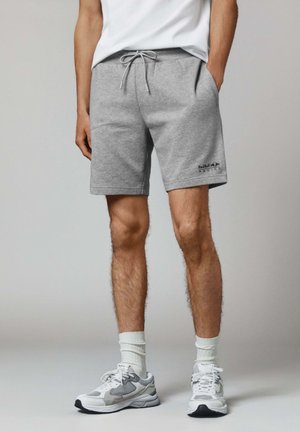 Pantalones cortos de algodón gris con cintura ajustable y bolsillos laterales. Presenta un logo de "Red Bull Racing" en el muslo izquierdo. Combinados con zapatillas blancas.