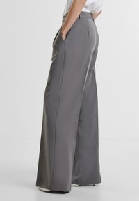 Šedé kalhoty s širokými nohavicemi a hladkým, matným povrchem, se bočními kapsami a přizpůsobeným střihem, stylizované s vysokým pasem a jemnými sklady.