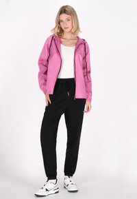 Giacca leggera rosa con cappuccio e dettagli neri, abbinata a pantaloni della tuta neri e sneakers bianche. Texture liscia, design sportivo.