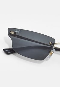Ray-Ban saulesbrilles ar elegantu melnu rāmjiem, pelēkiem stikliem un zelta detaļām. Iezīmējas ar izliekta stikla formu un skaidriem deguna spilventiņiem.