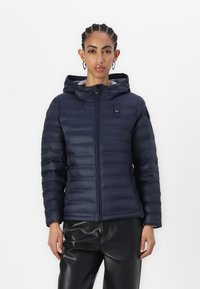 Marine blauwe donsjack met een capuchon, horizontaal gestikt ontwerp, voorkant met rits en logodetail. Draag het over zwarte leren broeken.