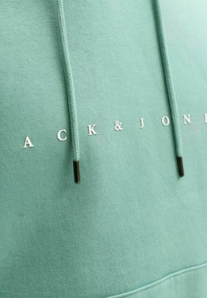 Piparmētru zaļš hoodijs, izgatavots no mīkstas auduma, ar baltām uzrakstām "ACK & JONES" un melno akcentu aparatūru uz stringa.