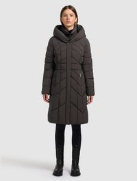 Mujer con un abrigo de invierno acolchado negro hasta la rodilla, con cuello alto, pantalones negros y botas negras, de pie frente a un fondo liso.