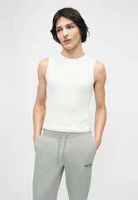 Νέος με λευκό αμάνικο τοπ και ανοιχτό γκρι φόρμα με τα χέρια στις τσέπες, μπροστά σε ένα απλό φόντο.