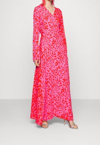 Langes pinkes Kleid mit rotbraunem Leopardenmuster, das ein Wickeldesign und lange Ärmel hat. Kombiniert mit klaren Absatzsandalen.