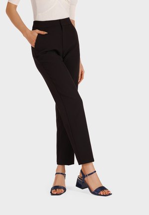 Pantaloni neri su misura con vestibilità slim e tasche laterali, abbinati a sandali blu con tacco a blocco e immancabili cinture con design testurizzato.