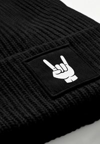 Black knit beanie featuring a rectangular black patch with a white hand gesture symbol, textured ribbed design throughout the fabric.