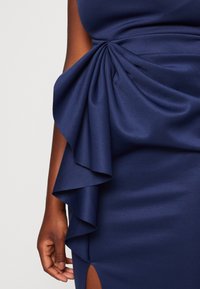 Robe bleu marine avec un tissu texturé, un détail de volants drapés sur le côté, et une fente haute sur la cuisse pour un intérêt de design supplémentaire.
