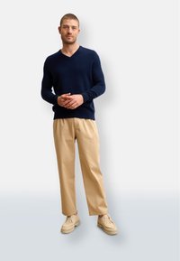 Marinblå stickad tröja med V-ringning, parat med beige vida byxor och ljusbruna snörskor, mot en neutral bakgrund.