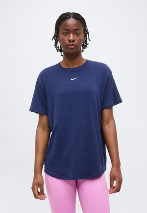 Γυναίκα με πλεξούδες στα μαλλιά, φορώντας μπλε σκούρο t-shirt της Nike και ανοιχτό ροζ κολάν, στέκεται μπροστά σε ένα απλό φωτεινό φόντο.