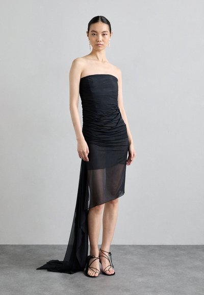 Μαύρο strapless φόρεμα με ρυάκινο ύφασμα, ασύμμετρο τελειώμα και διάφανο επικάλυμμα. Φοριέται με μαύρες σανδάλια με λεπτές λωρίδες, που προβάλλουν επίπεδες σόλες.