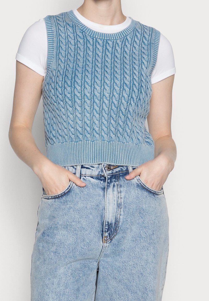 Pull sans manches en bleu clair avec un design en tricot torsadé, associé à un t-shirt blanc et un jean bleu clair taille haute.