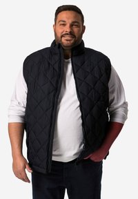 Černá prošívaná vesta s vysokým límcem, zipovým zavíráním a diamantovými vzory, nošená přes bílou košili, spárovaná s tmavými džíny.