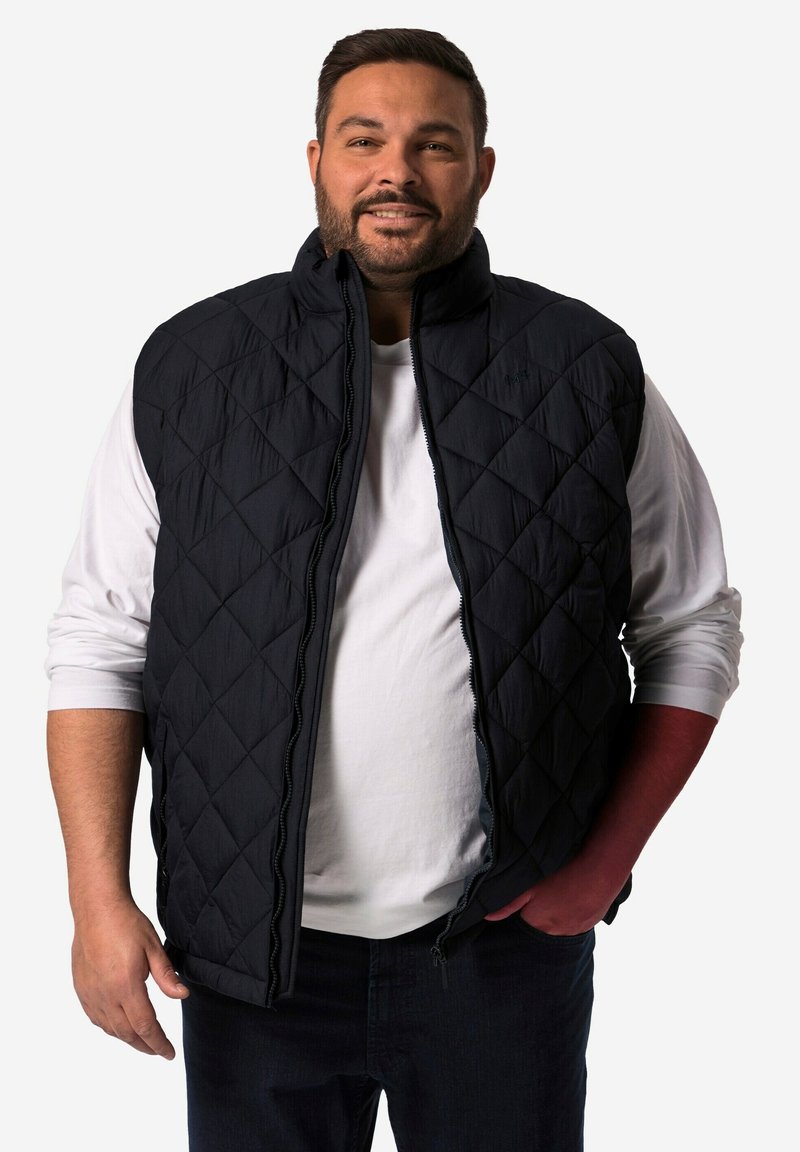 Černá prošívaná vesta s vysokým límcem, zipovým zavíráním a diamantovými vzory, nošená přes bílou košili, spárovaná s tmavými džíny.