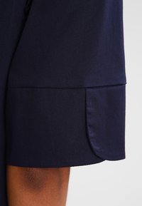 Mörkblå ärmsydd med en jämn textur, vikbar manschett med rundade kanter och subtil sömdekoration längs sömmen.