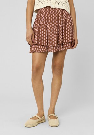 Femme portant une mini-jupe fleurie marron, un top beige en crochet et des espadrilles beiges, debout devant un fond uni.