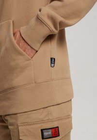 Beżowy sweter i spodnie cargo z czarnymi i czerwonymi łatami Dakar, ręka wsunięta w kieszeń swetra.