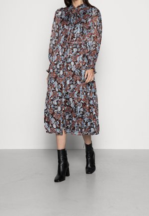 Femme portant une robe à manches longues, de longueur moyenne, à motifs floraux dans des tons bleus, bruns et blancs, associée à des bottines noires à talons carrés.