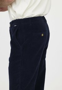 Marineblaue Cordhosen mit vertikalen Streifen, ausgestattet mit einer Gesäßtasche und einem Goldknopf-Detail am Bund. Glatte Textur.