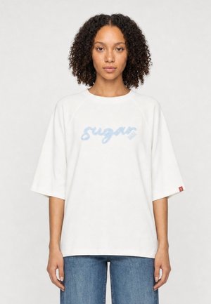 Woman with curly hair wears a white oversized shirt with blue "sugar" text and blue jeans against a plain light background.