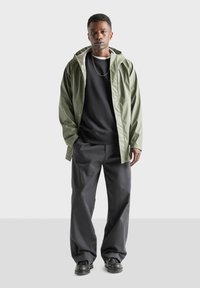 Olivgrön vattentålig jacka över en svart sweatshirt, tillsammans med lösa svarta byxor och svarta skor, med en enkel design och neutrala toner.