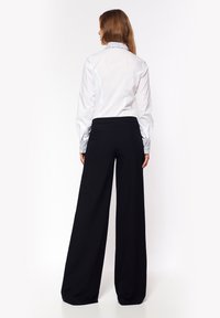 Kobieta z długimi brązowymi włosami stoi tyłem, ubrana w białą koszulę z wzorzystymi mankietami i kołnierzykiem oraz szerokimi czarnymi spodniami.