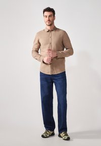 Camicia button-up marrone chiaro con maniche lunghe, abbinata a jeans dritti blu scuro e sneakers con motivo camouflage verde.