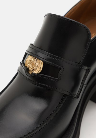 Schwarze Lederloafer mit hochglänzender Oberfläche, ausgestattet mit einem dekorativen goldenen Emblem und kontrastierenden Nähten entlang der Kanten.